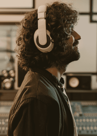 Young man with dark curly hair and white headphones on, listening to music in a studio environment with his eyes closed