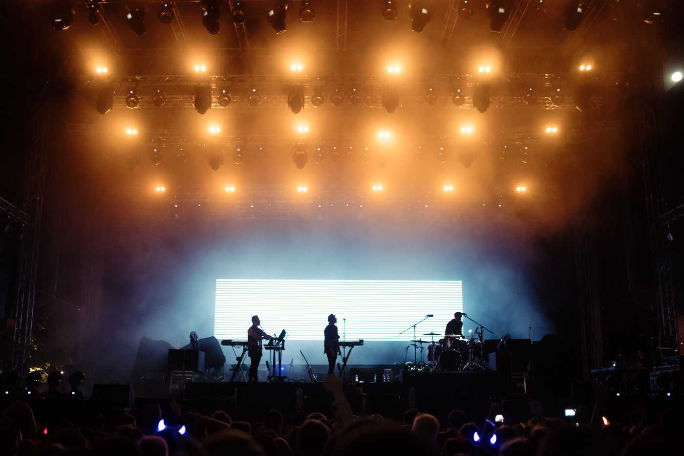 Image of a concert hall with big bright orange lights on the ceiling and on stage playing a three man band.